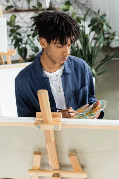 Молодий афроамериканець, який змішує фарби на палітрі біля полотна. — стокове фото