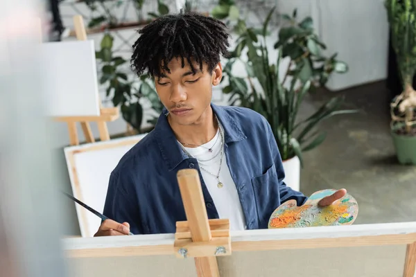 Jovem artista afro-americano pintando sobre tela e segurando paleta com tintas em estúdio — Fotografia de Stock