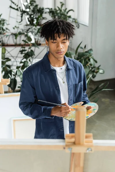 African american artist holding paintbrush and palette near blurred easel and canvas — Stock Photo