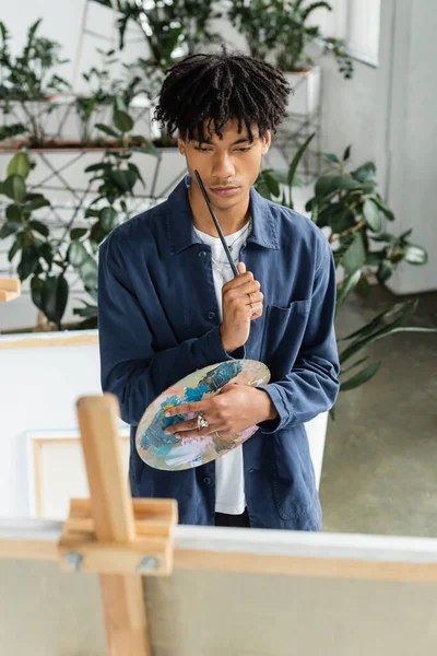 Focused african american artist holding paintbrush and palette near canvas — Stock Photo