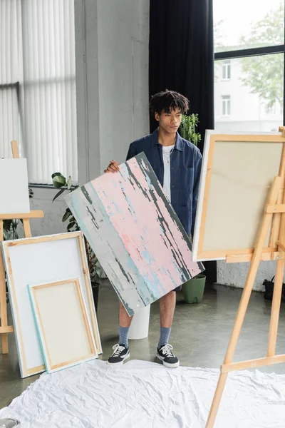 Young african american artist holding painting near canvas on easel in studio — Stock Photo