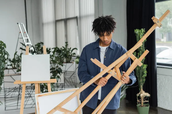 Young african american artist holding wooden easel in workshop — Stockfoto