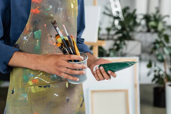 Cropped view of african american artist in apron holding paintbrushes and paint tube — Stock Photo