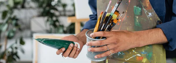 Cropped view of afrcan american artist in apron holding paintbrushes and paint, banner — Stock Photo