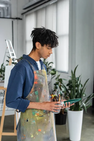 Вид сбоку на молодого африканского американского художника в фартуке, держащего кисти и краски в студии — стоковое фото