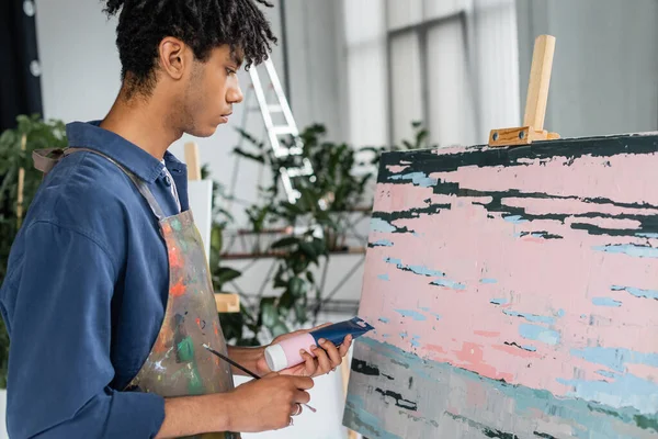 Side view of african american artist holding paint and paintbrush near abstract painting in studio — Stock Photo