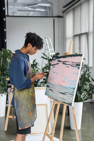 Side view of african american artist in apron holding paintbrush and paint near drawing in studio — Stock Photo