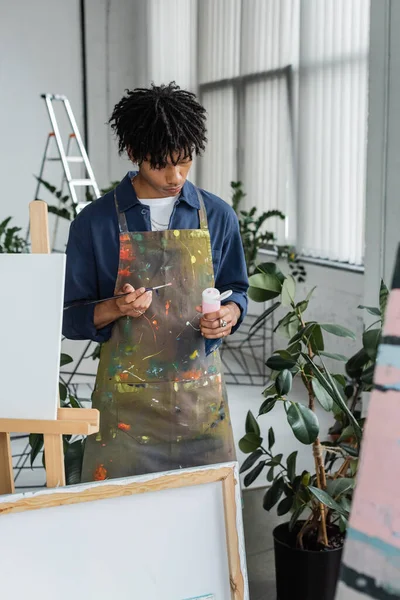 African american artist in apron holding paintbrush and paint near canvases in workshop — Fotografia de Stock