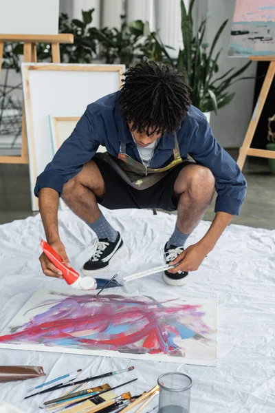 African american artist pouring paint on canvas near paintbrushes on cloth in studio — Stock Photo