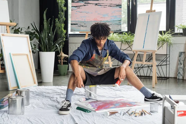 Young african american artist holding paint and paintbrush near canvas on floor in workshop - foto de stock