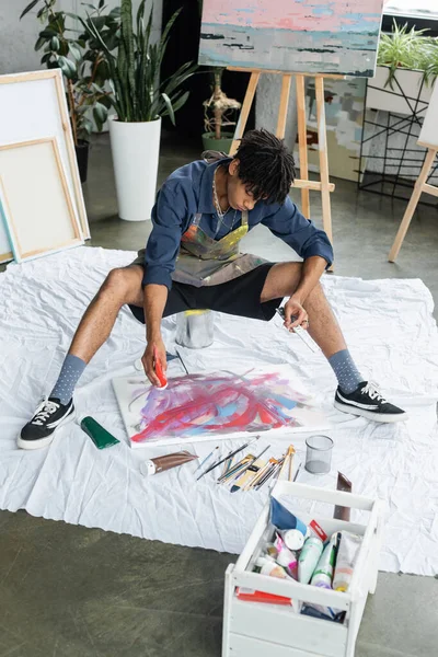 Young african american artist pouring paint on canvas near paints in workshop - foto de stock