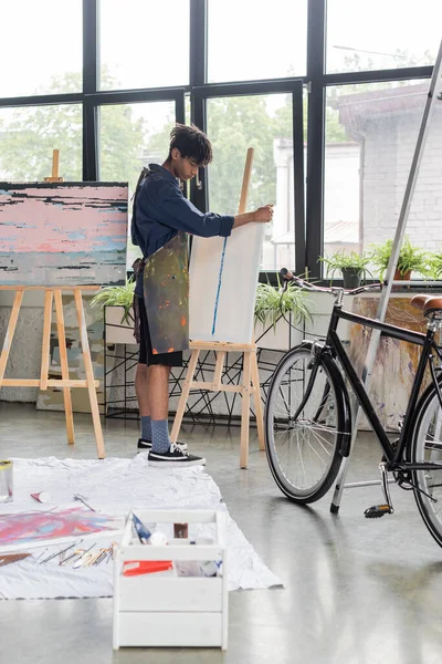 Side view of young african american artist in apron putting canvas on easel near bike in workshop — Stock Photo