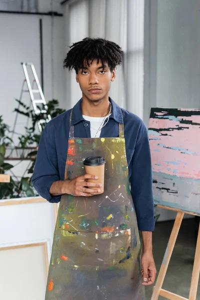 African american artist in apron holding coffee to go in studio - foto de stock