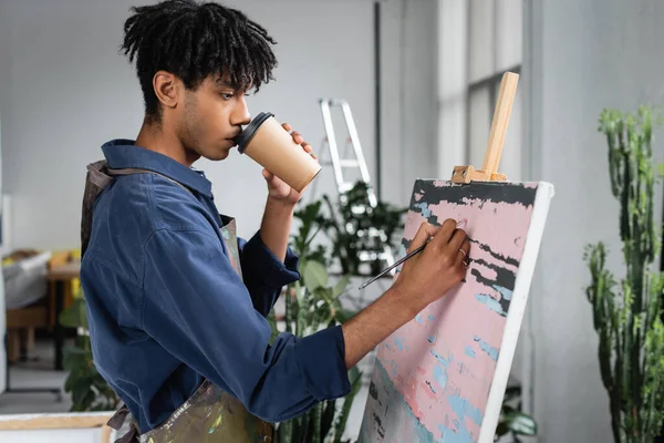 Young african american artist drinking coffee to go and painting on canvas in workshop — Stock Photo