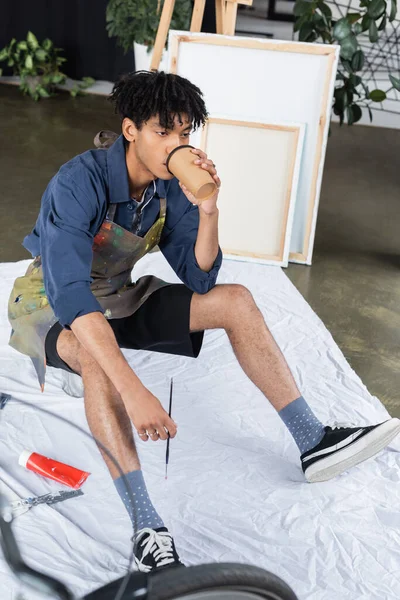 African american artist in apron drinking coffee to go and holding paintbrush in studio — Stock Photo
