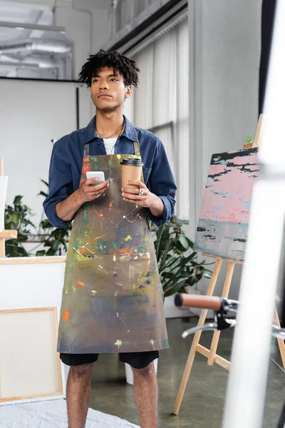 Pensive african american artist in apron holding smartphone and coffee in workshop — Stock Photo