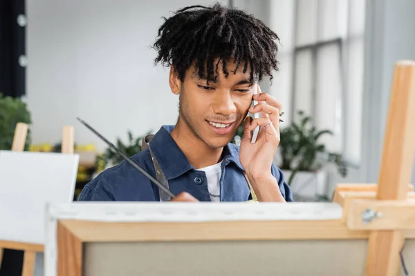 Smiling african american artist talking on smartphone and painting on canvas in studio — Stock Photo