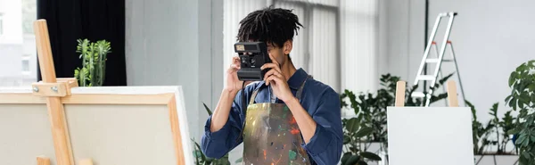 Artista afroamericano che scatta foto su fotocamera vintage vicino a tela su cavalletto in studio, banner — Foto stock