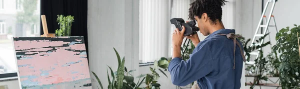 Young african american artist taking photo on retro camera near drawing in workshop, banner — Stock Photo