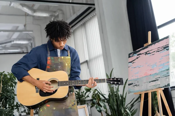 Young african american artist in apron playing acoustic guitar in workshop — Stock Photo