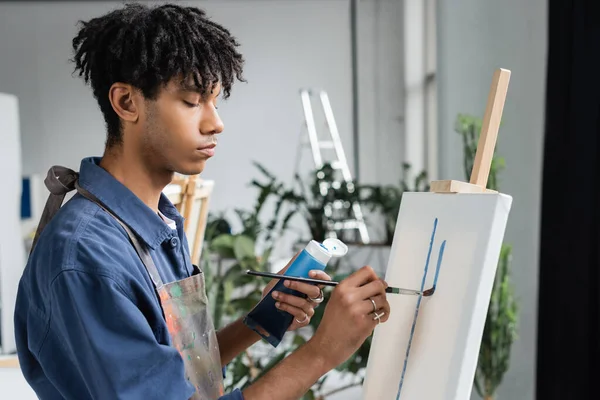 African american artist in apron painting on white canvas in studio — Fotografia de Stock