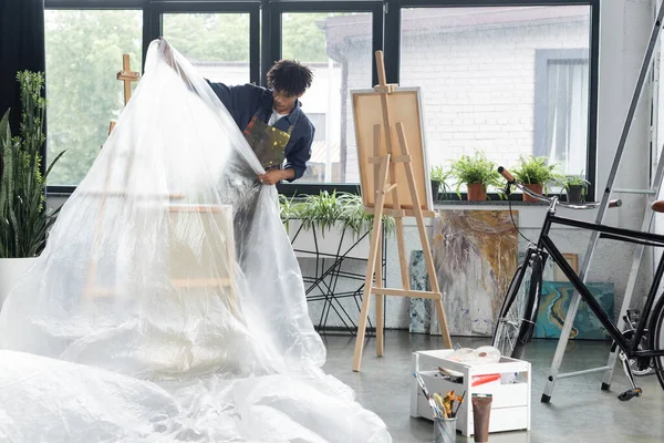 African american artist covering easel and canvases with cellophane in workshop — Stock Photo