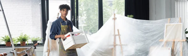 Африканський американський художник у фартусі тримає коробку біля мольберта в целофані в студії, банер — стокове фото