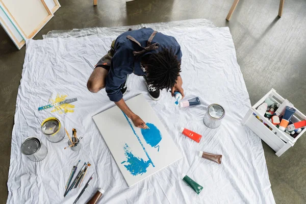 Overhead view of african american artist painting on canvas near paints on floor in studio — Stock Photo