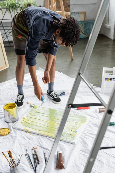 African american artist holding acrylic paint near canvas in workshop - foto de stock