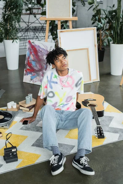 Joven artista afroamericano sentado cerca de smartphone y guitarra acústica en estudio - foto de stock