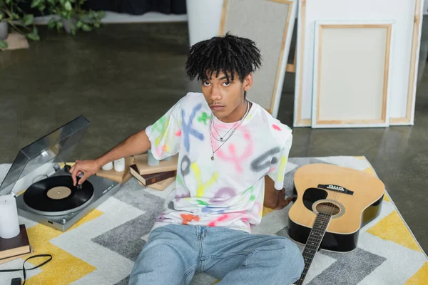 Young african american artist using record player near acoustic guitar and canvases in studio — Fotografia de Stock