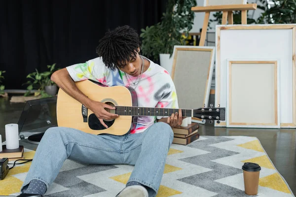 African american artist playing acoustic guitar near canvases and coffee in studio — Stock Photo