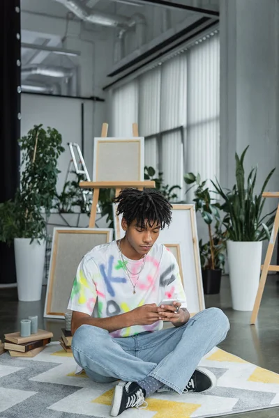 Молодий афроамериканець, який використовує смартфон поблизу книг і полотен у художній студії. — стокове фото