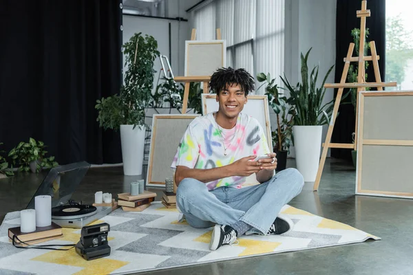 Усміхнений афроамериканський художник використовує смартфон поблизу старовинної камери і звукорежисера в майстерні — стокове фото