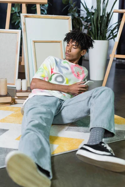 Jeune artiste afro-américain tenant un smartphone près de toiles et de livres en studio — Photo de stock