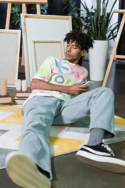 Young african american artist holding smartphone near canvases and books in studio — Stock Photo