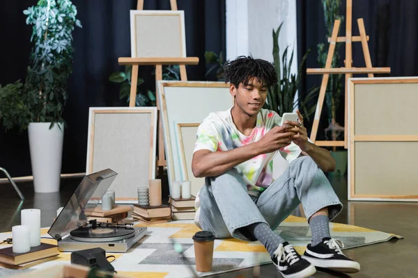 Усміхнений афроамериканський художник, використовуючи смартфон біля кави, щоб піти і полотна в студії — стокове фото