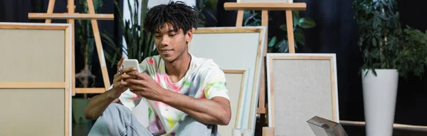 Positive african american artist using smartphone near canvases in workshop, banner - foto de stock