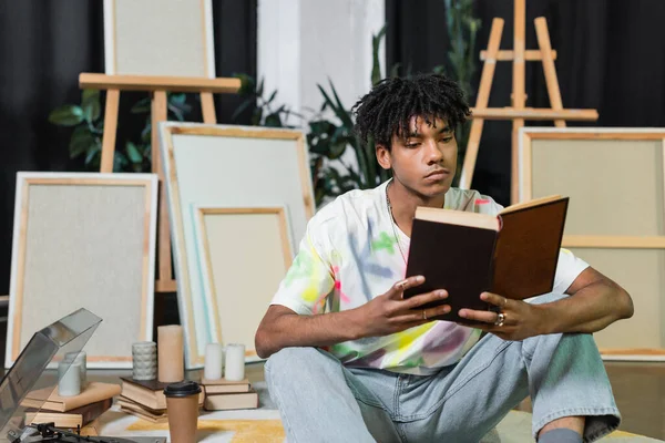 African american artist reading book near record player and canvases in workshop — Stockfoto