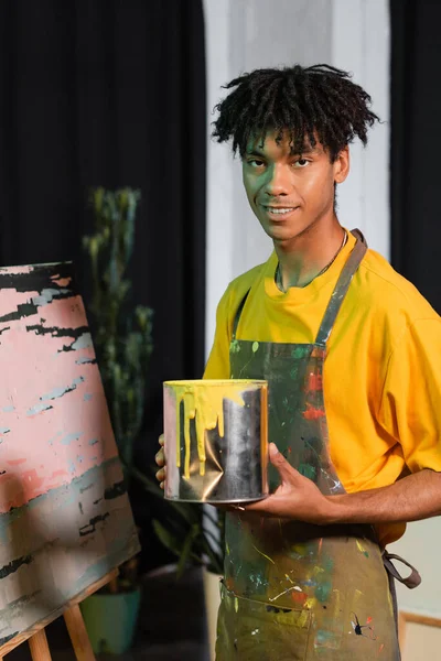 Smiling african american artist in apron holding jar with paint near painting in studio — Stock Photo
