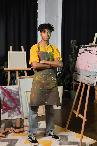 Sonriente artista afroamericano cruzando brazos cerca de pinturas en estudio - foto de stock