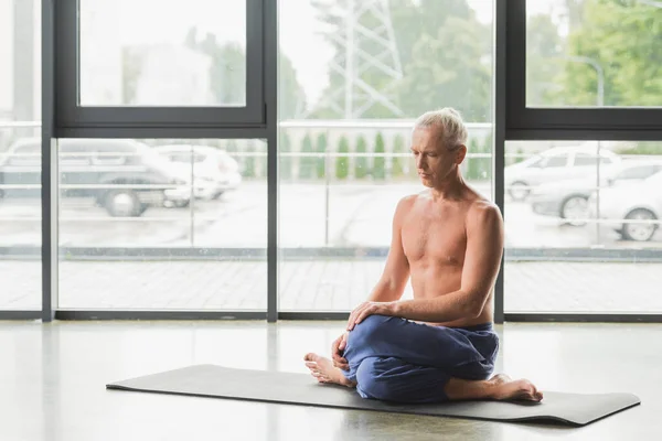 Hombre descalzo en pantalones azules sentado en la postura de yoga torcido en la estera - foto de stock