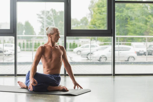 Grey haired man in blue pants sitting in twisting yoga pose on mat in studio — Fotografia de Stock