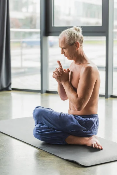 Hombre de pelo gris en pantalones sentados en pose de yoga retorcido y haciendo chakra mudra corona - foto de stock