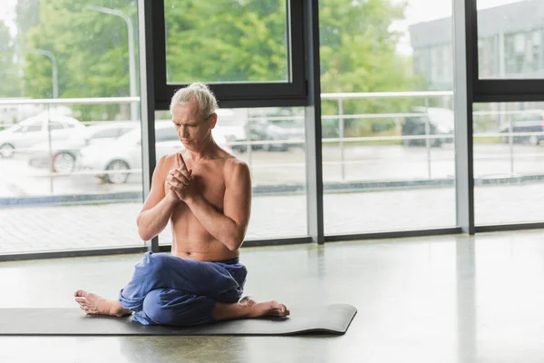 Hombre de pelo gris en pantalones sentados en pose de yoga retorcido y haciendo chakra mudra corona cerca de ventanas - foto de stock