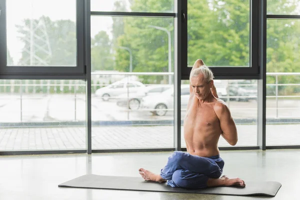 Мужчина без рубашки в синих штанах сидит в позе йоги и растягивается назад — стоковое фото