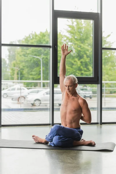 Hombre sin camisa en pantalones azules sentado en la postura de yoga torcido con el brazo levantado - foto de stock
