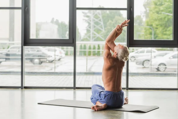 Shirtless man in blue pants sitting in twisting yoga pose and raising arms while stretching back — Stock Photo