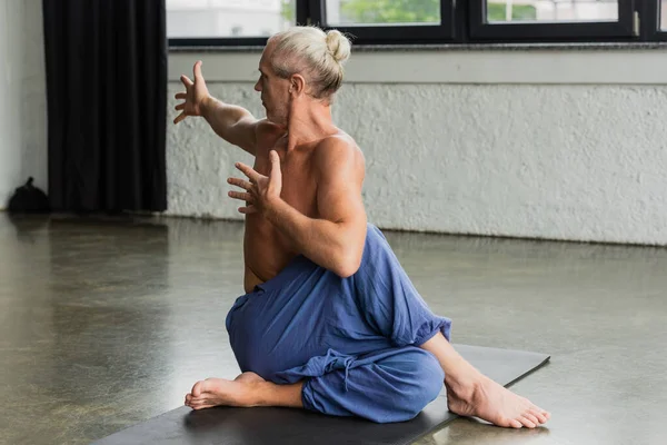 Homme aux cheveux gris en pantalon assis en demi-seigneur de poissons pose en studio de yoga — Photo de stock