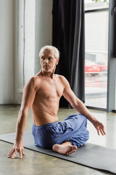 Shirtless and grey haired man in pants sitting in half lord of fishes pose in yoga studio — Photo de stock